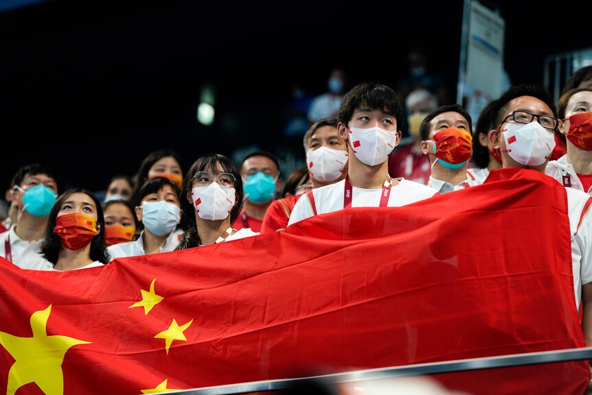 Women and men wearing face masks hold up a Chinese flag, standing shoulder to shoulder to spectace a sports event from a stand.