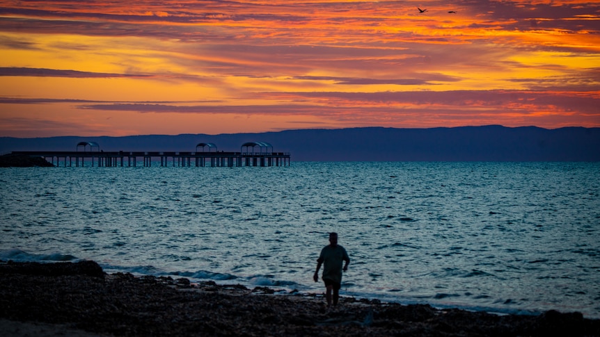 A person walks along the shoreline at sunset.