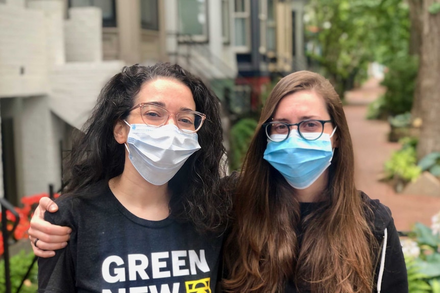 Two young women in casual clothes and face masks put arms around in each other in leafy street.