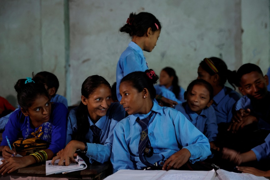 A group of teen girls crowd around an older woman, all wearing the same blue uniform, with schoolbooks in front of them 