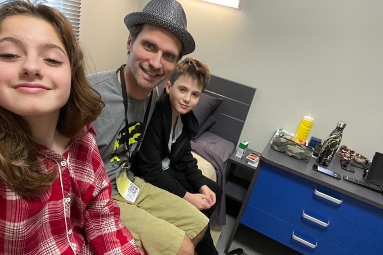 Three people sit on the edge of a single bed in a small room with a set of drawers.