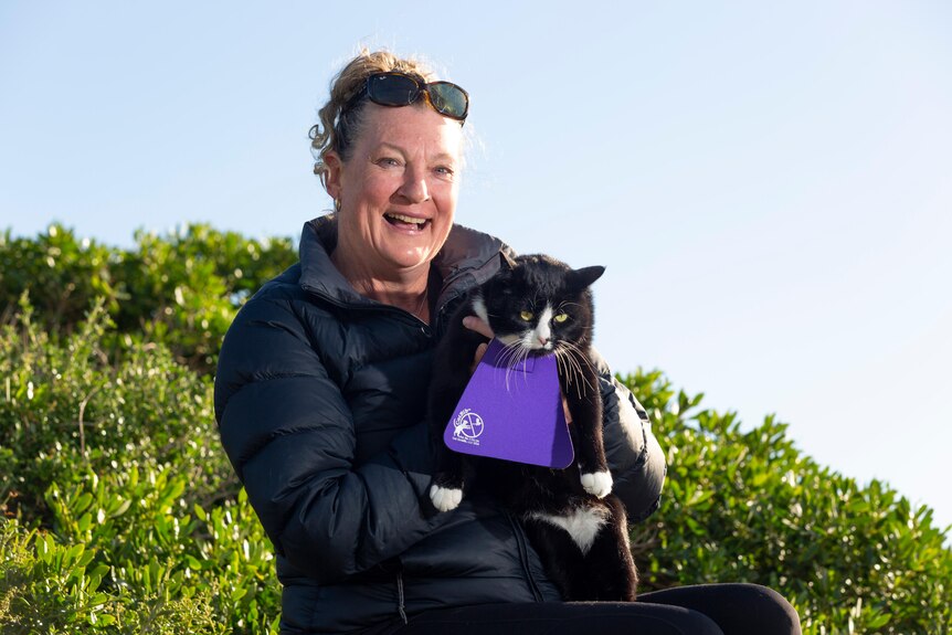 A woman standing outside holding a black and white cat that is wearing a bib.