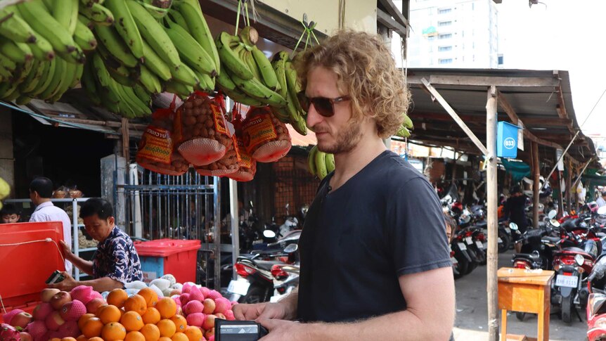 A man with curly hair and wearing sunglasses buys fresh fruit from a Cambodian market stall