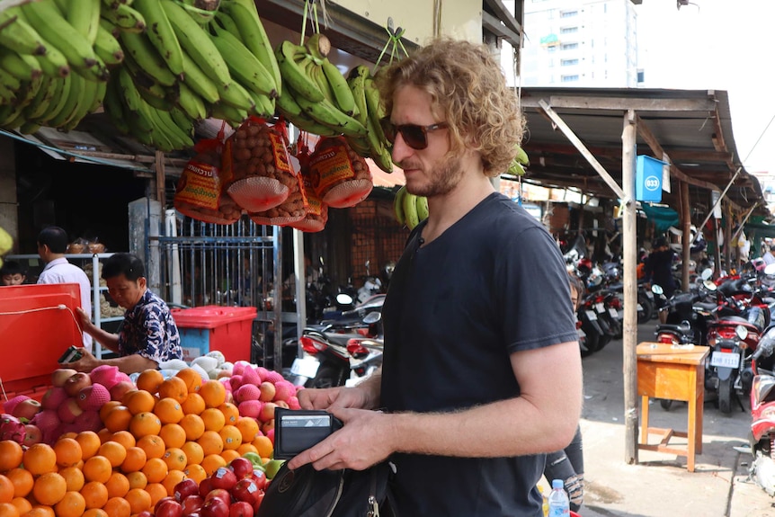 A man with curly hair and wearing sunglasses buys fresh fruit from a Cambodian market stall