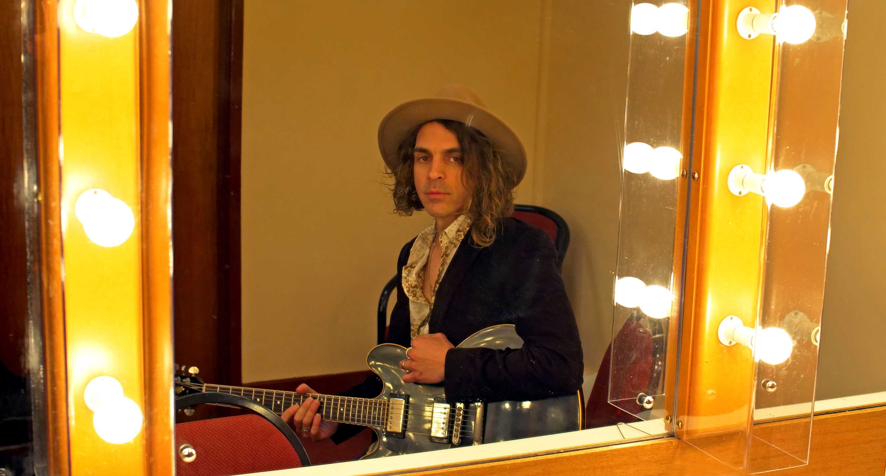 A portrait of a man holding a guitar and wearing a wide brim hat reflected in a mirror that's lined with light bulbs