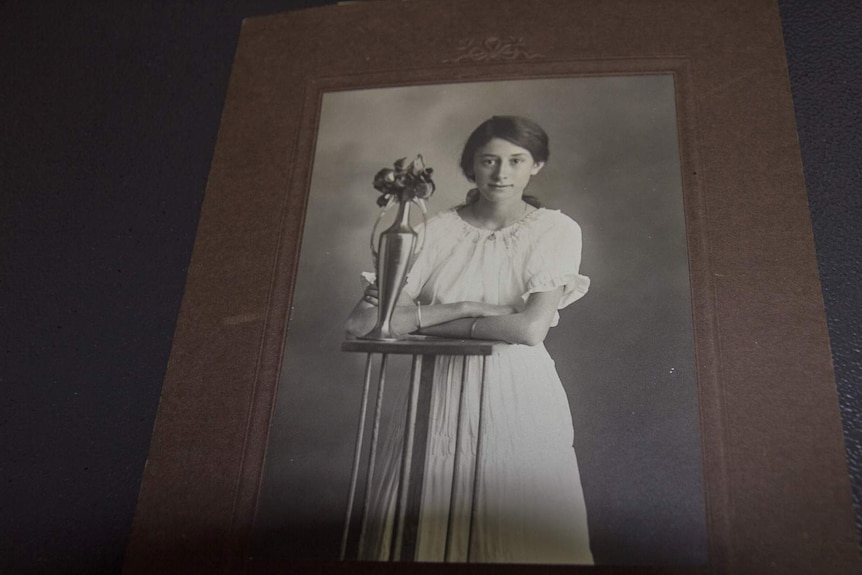 A black and white photograph in a brown cardboard frame of a teenaged girl in a white dress standing at a table with a vase