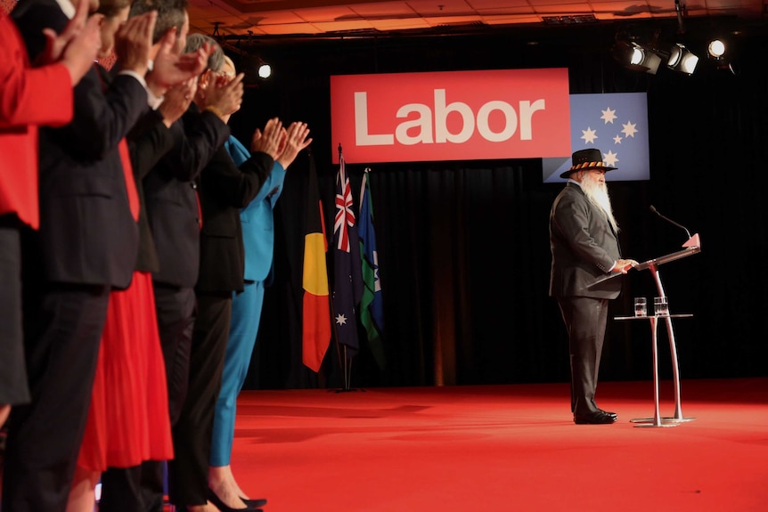 Pat Dodson stands a t a podium with Labor branding above him and flags behind him while receiving a standing ovation.