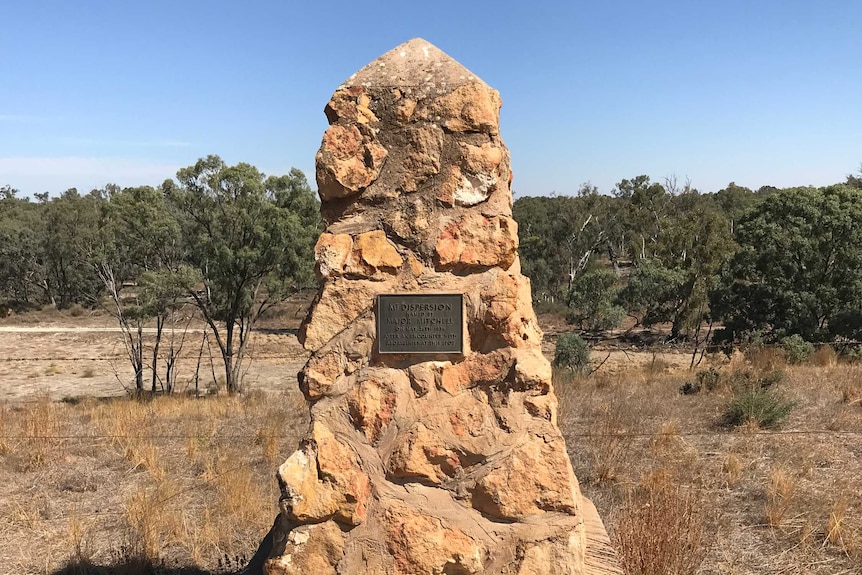 A stone cairn on dry grass surrounded by bushland with a blue sky and trees in the background.