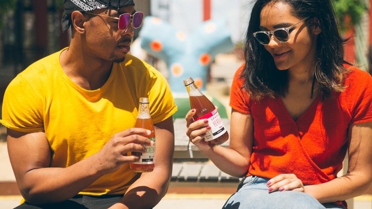 A young man in a yellow tshirt and sunglasses and a young woman with dark hair wearing a red tshirt tap their drinks