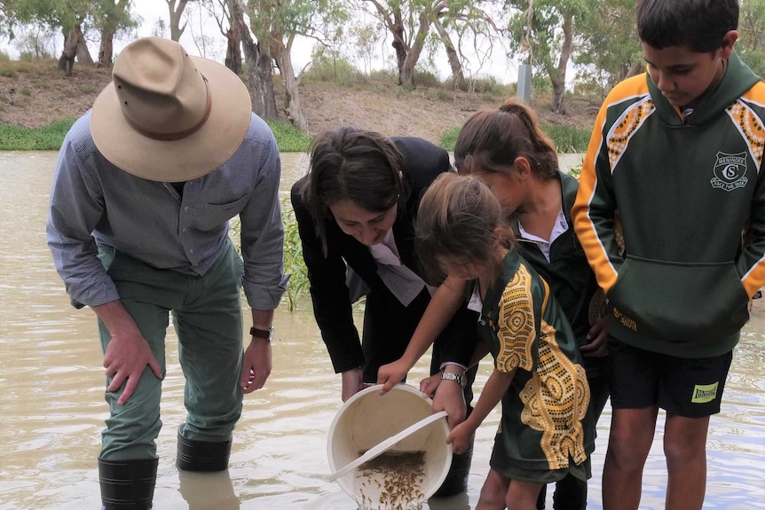 Two adults watch some school kids pour a bucket of small fish into a river.