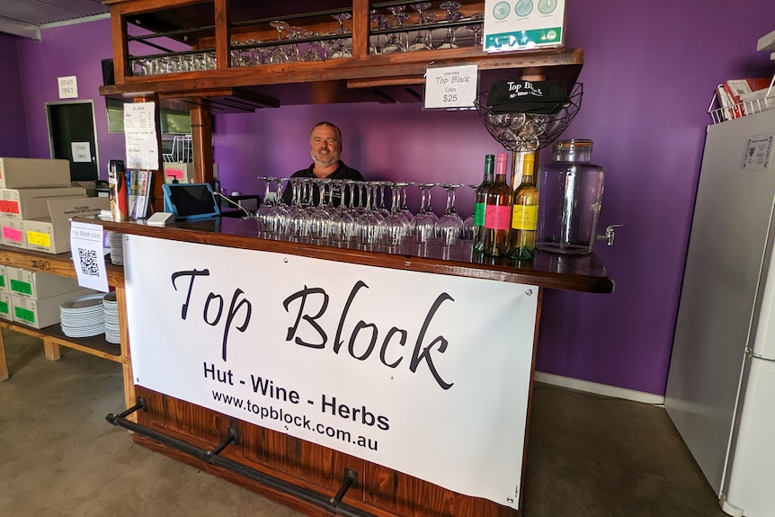 Mr Heward stands behind a wooden bar, with wine glasses and wine bottles in front a purple wall. 