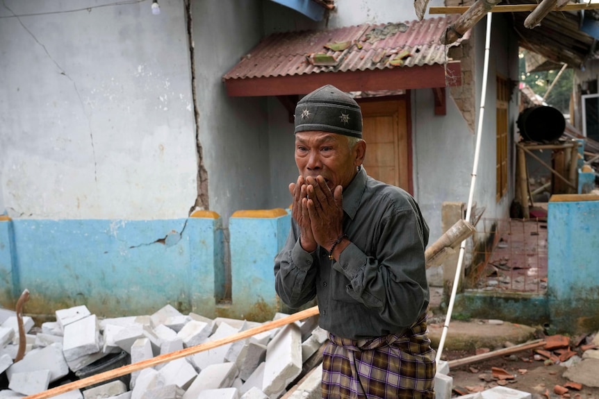 A man stands with his hands over his mouth amidst a damaged village. 