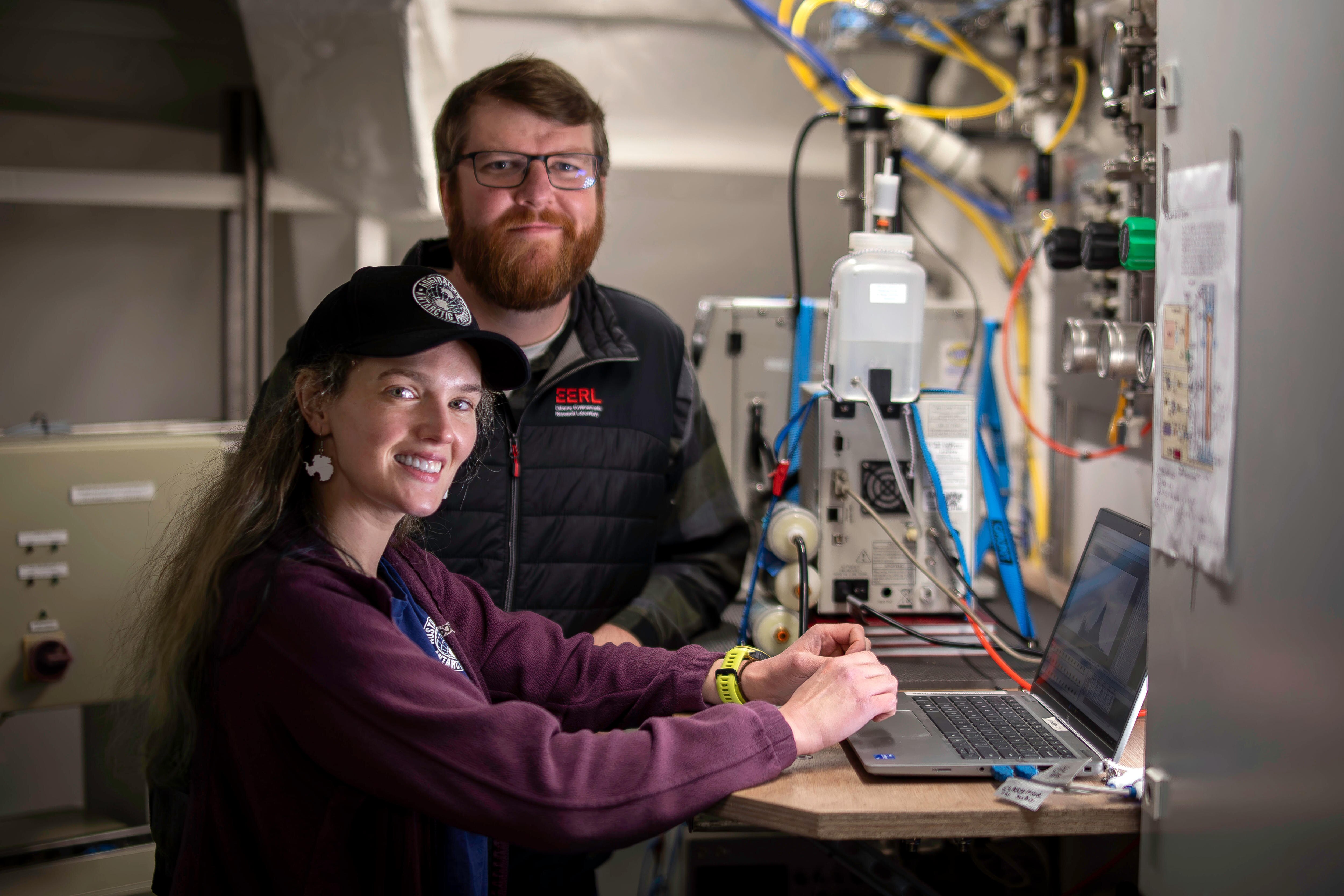 A woman in a purple jumper and black cap sits in front of a laptop computer. A man in black jumper stands behind her.