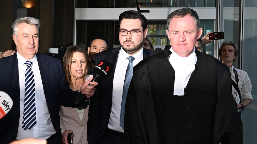 A man walks out of glass doors flanked by a lawyer and journalists.