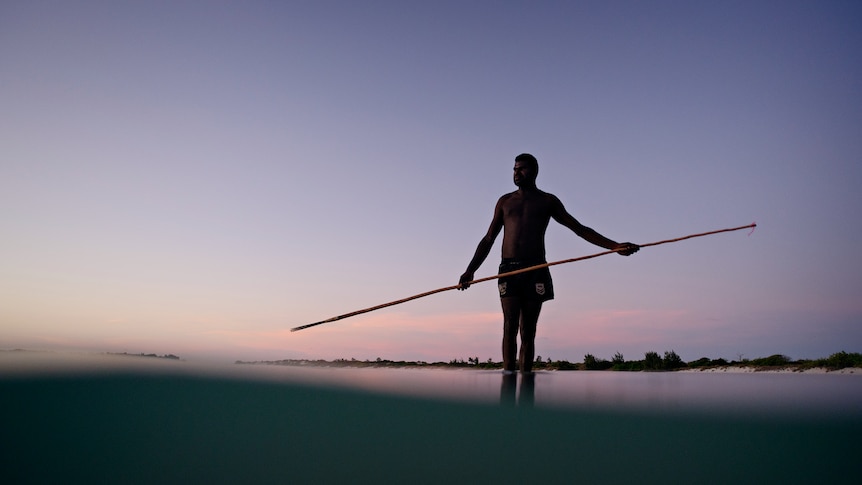A man standing in the water off a beach and holding a long spear, at sunset