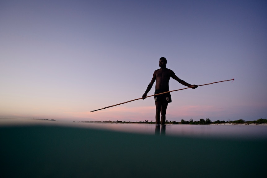 A man standing in the water off a beach and holding a long spear, at sunset