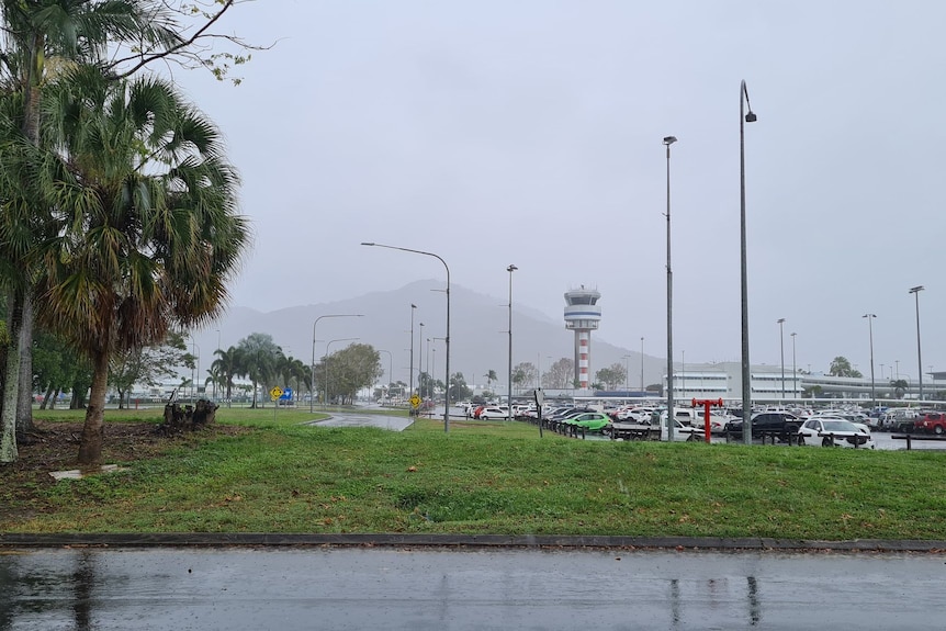 Grey sky, rain and fog covers Cairns, wet road and trees in foreground, shopping centre in background.