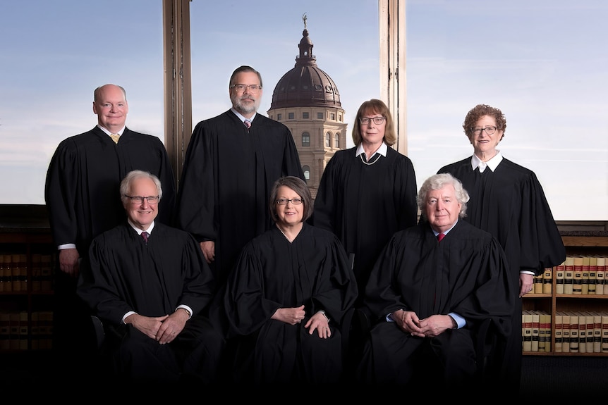 Kansas's Seven Supreme Court Justices pose in a portrait photo with the Kansas Supreme Court visible in the background