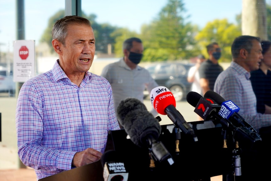 A wide shot of Roger Cook standing in front of a bank of microphones.