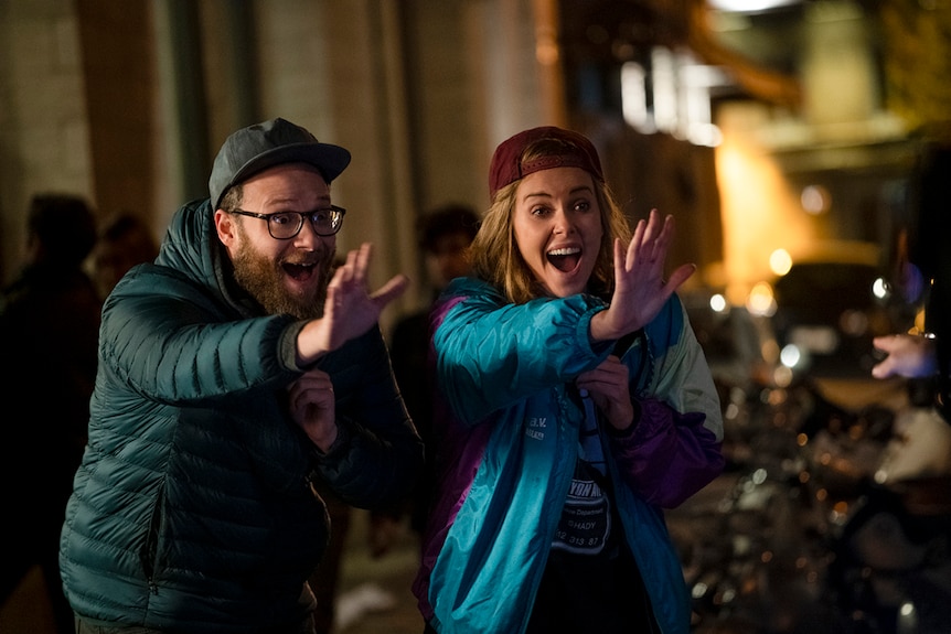 Colour still of Seth Rogen and Charlize Theron wearing caps, arms extended and yelling at night time in 2019 film Long Shot.