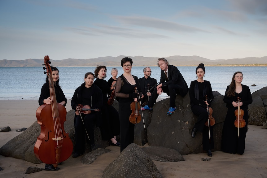 A nine piece classical music ensemble poses on a beach against a mountain backdrop.