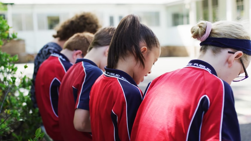 Several young children wearing red and blue shirts site on a wall in a courtyard.