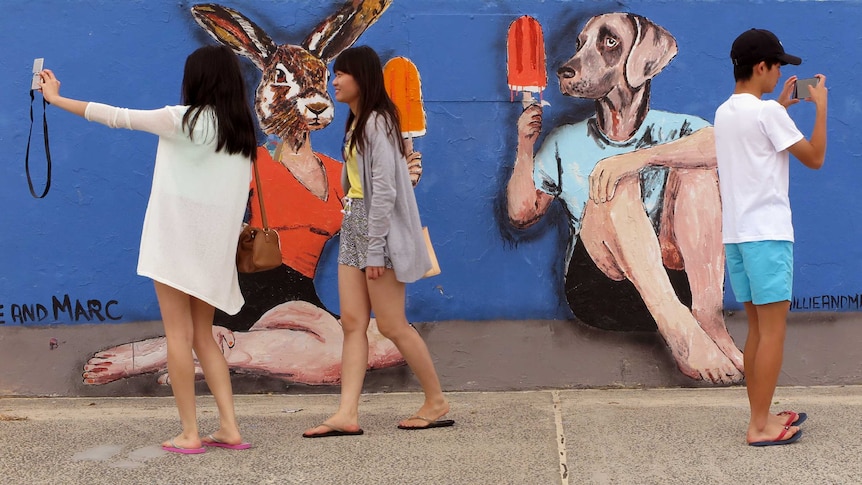 Three young people in shorts take selfies in front of a street painting of a body with a rabbit head and one with a dog head.