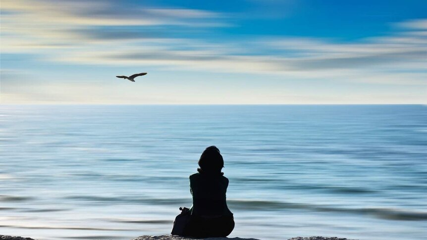 A woman in shadow sits on a rock and looks at a serene, calm ocean.