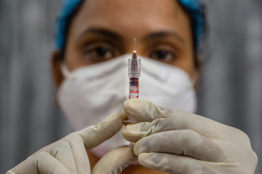 A woman wearing a mask and gloves holds a syringe in front of her face.