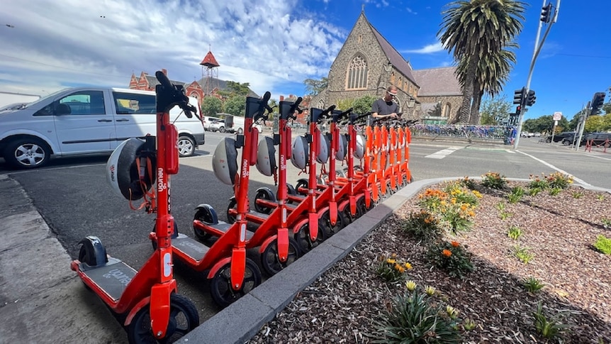 A dozen orange e-scooters lined up along a Ballarat street, with a church and road in the background.