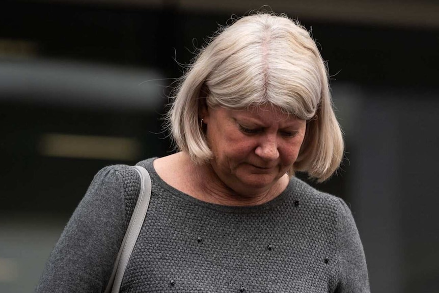 A woman with shoulder-length grey hair and wearing a dark grey jumper in front of a court building.