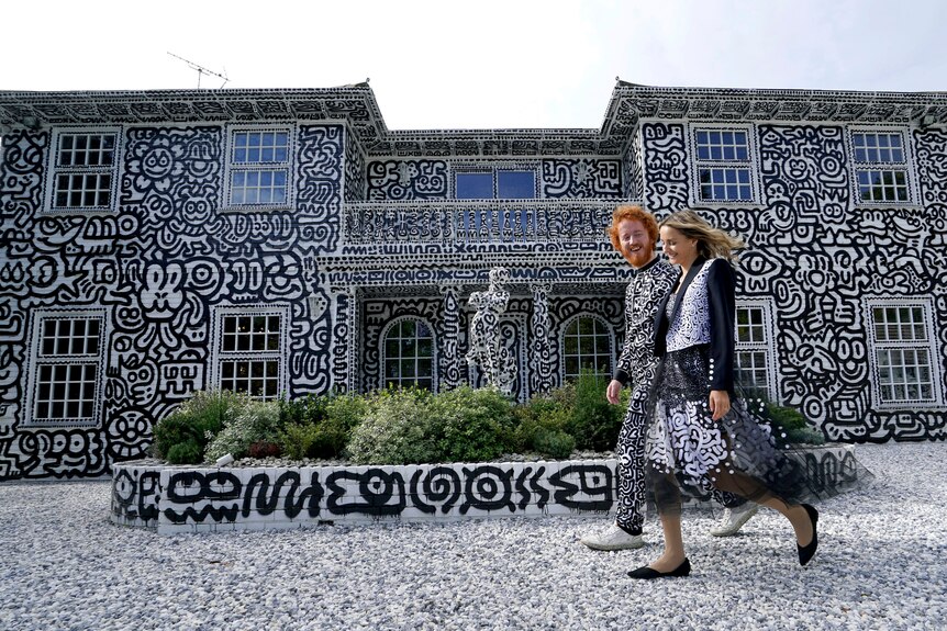 Artist Sam Cox and his wife walk past a two-storey home and garden all covered in black and white art. 