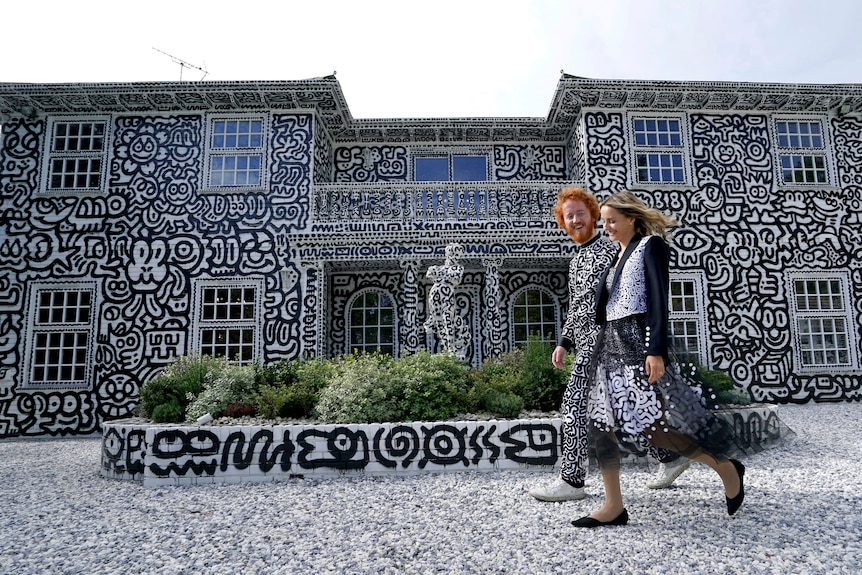 Artist Sam Cox and his wife walk past a two-storey home and garden all covered in black and white art. 