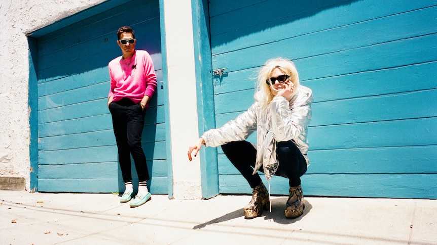 Jamie Hince and Alison Mosshart of The Kills stand in front of a bright blue garage door.