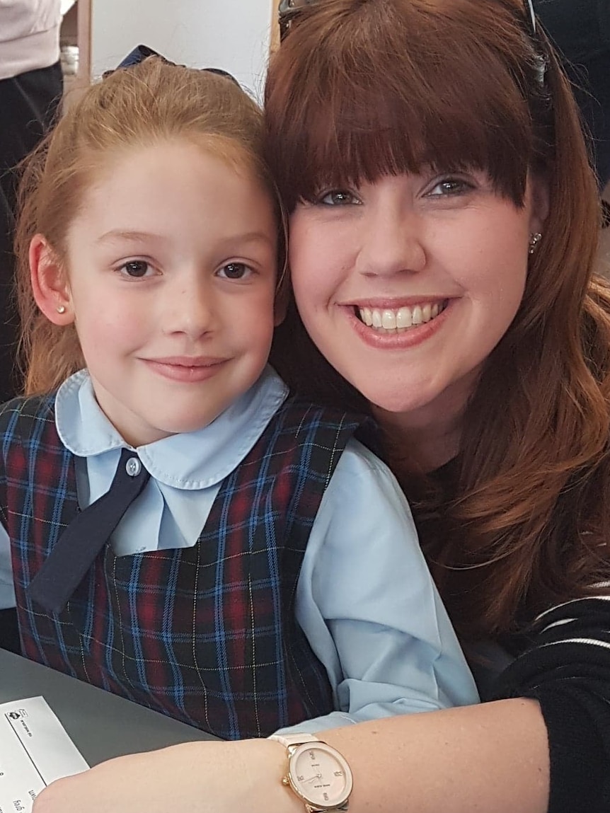 Mother sits with daughter who is wearing school uniform at a school desk.