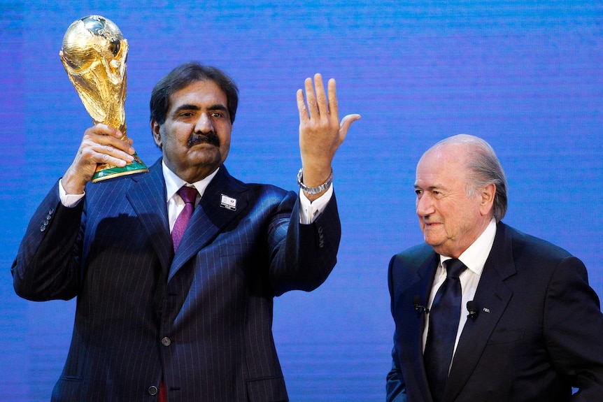 A man in a pinstripe suit holds up a gold trophy in one hand while gesturing in the other, while another man in a suit watches