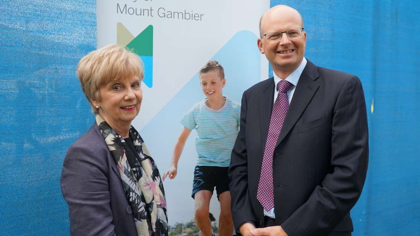 A blonde haired woman and a balding man with glasses stand in front of a poster reading "City of Mount Gambier"