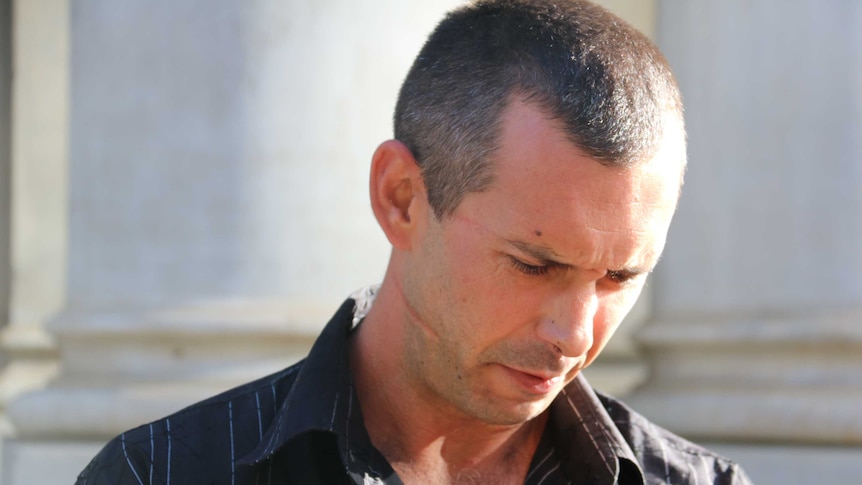 A man with two mid-size scars on one side of his face looks down at the ground outside court.
