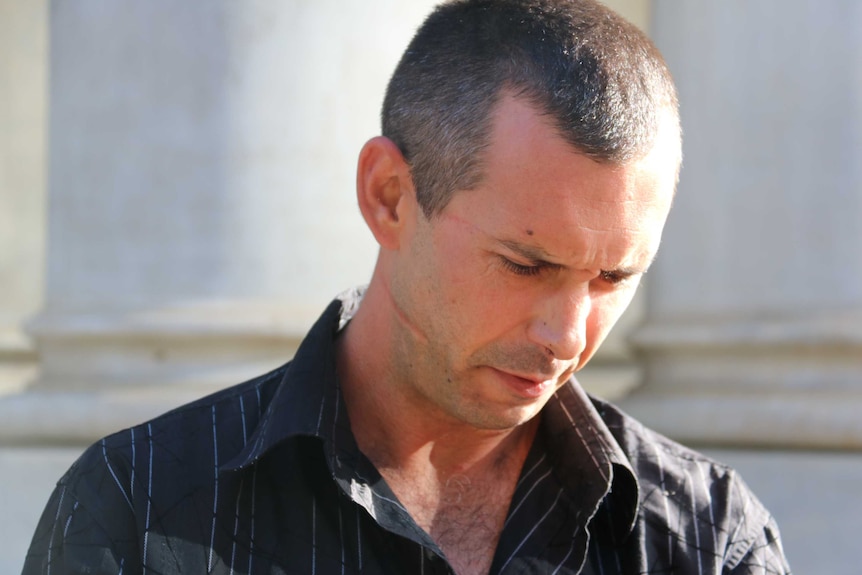 A man with two mid-size scars on one side of his face looks down at the ground outside court.