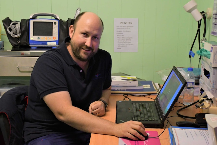 A bald man with a daqrk blue shirt sits at a desk and looks at the camera.