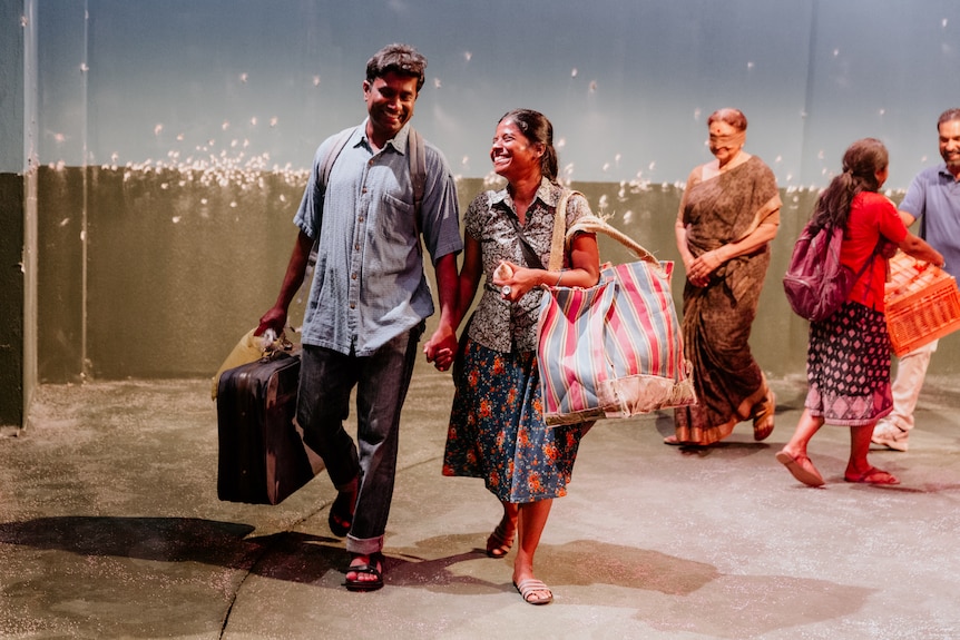 A young Sri Lankan man and woman holding baggage and each other's hands walk on a stage, older Sri Lankan woman in background