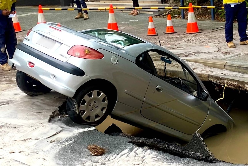 A silver car is partially submerged in a sinkhole in the road