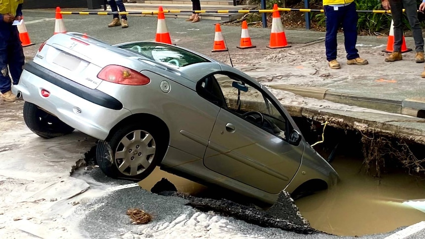 A silver car is partially submerged in a sinkhole in the road