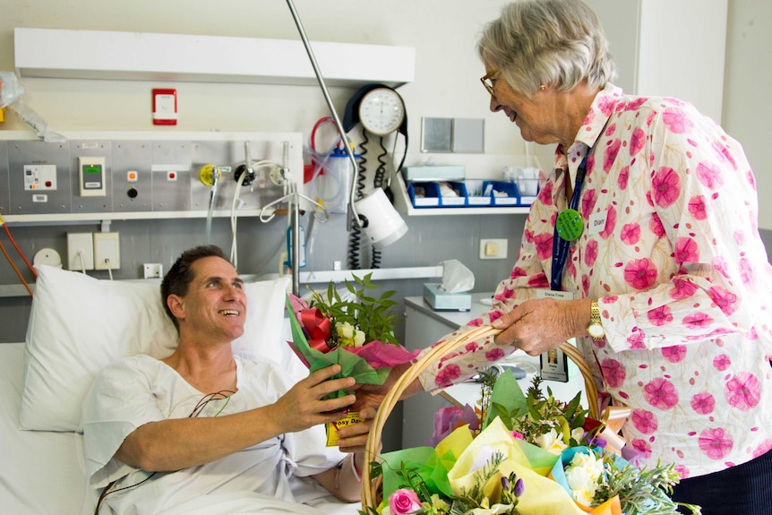 A man lying in a hospital bed smiles as a woman hands him a bunch of flowers.