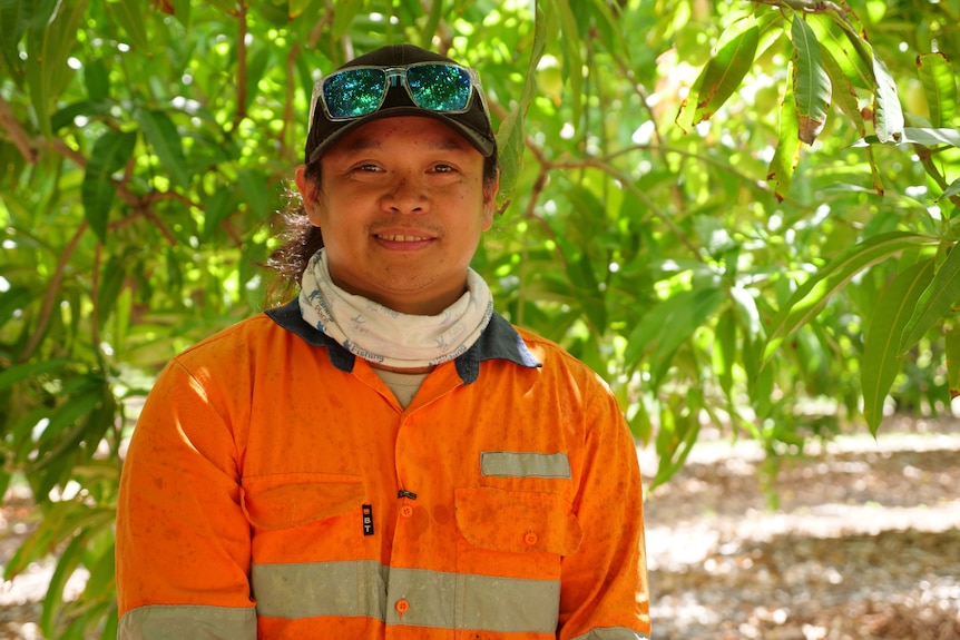 A young man smiles shaded under a mango tree. He's wearing high-vis work gear is using a scarf and cap as sun protection