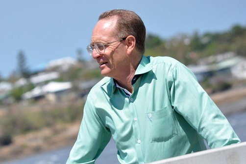 A bespectacled man stands outside, squinting in the sun.