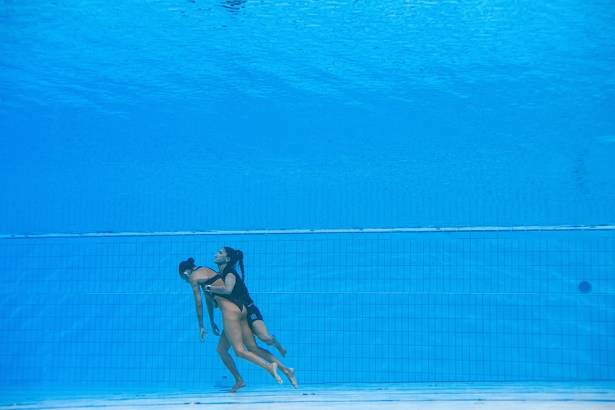 A woman, carrying an unconscious woman, swims from the bottom of a pool to the surface.