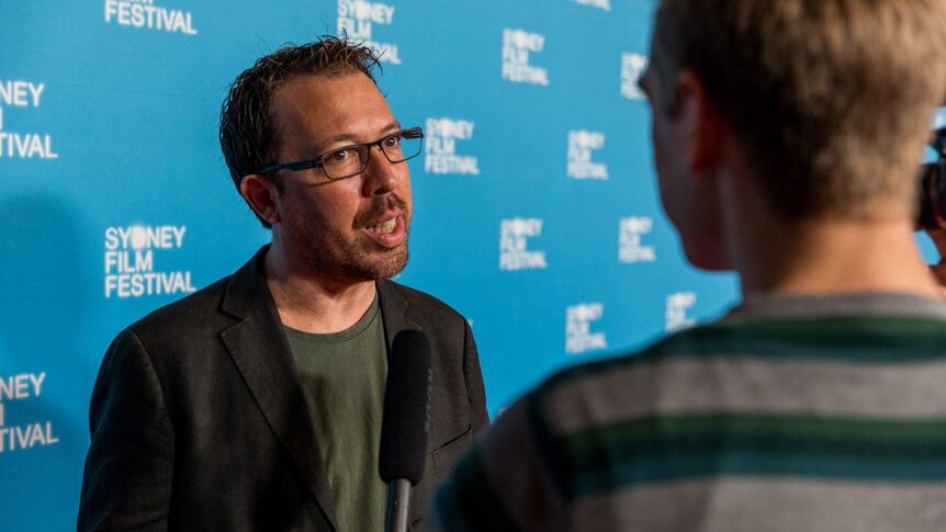 Director has short hair, rough shaven, wearing dark-rimmed glasses, dark jacket and tee standing against SFF red carpet wall.