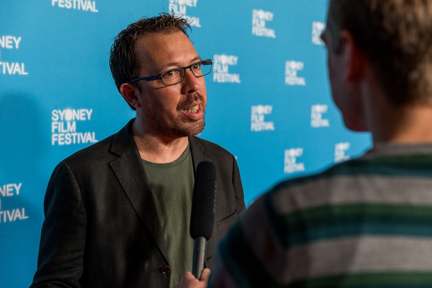 Director has short hair, rough shaven, wearing dark-rimmed glasses, dark jacket and tee standing against SFF red carpet wall.