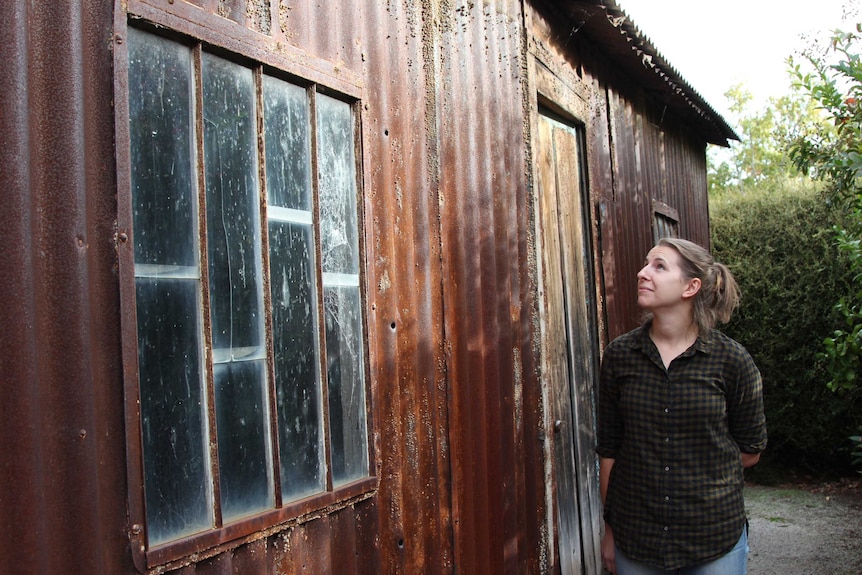 National Trust assets coordinator Alyce McCue looks up at the exterior of a brown corrugated iron home.